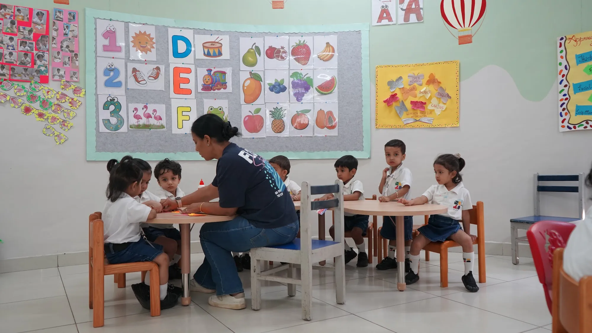 A teacher sitting with a small group at their table, a fruit and vegetable chart on the wall behind them