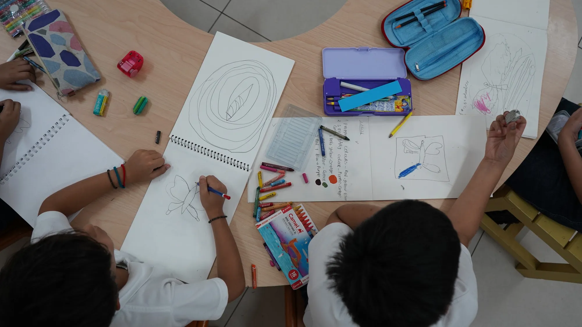 Children writing and drawing on sheets of paper around a classroom table, seen from above